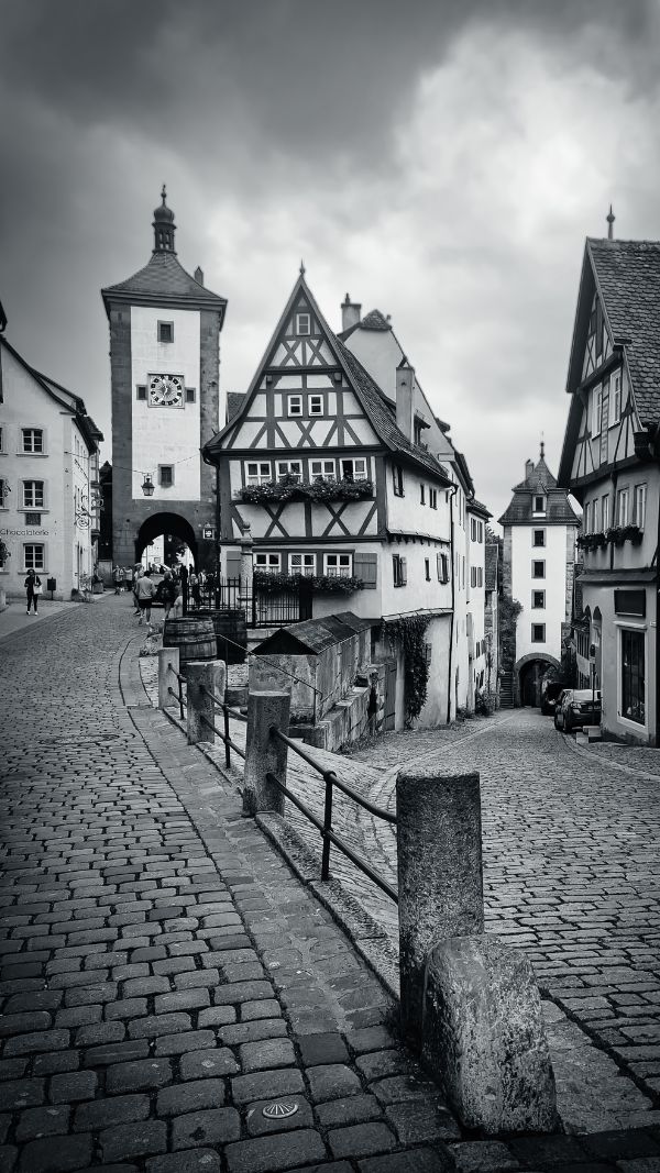 Cobblestone street leading to a historic clock tower flanked by traditional half-timbered houses under a cloudy sky.