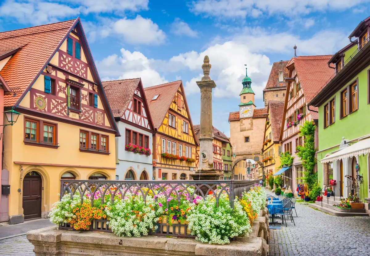 A charming street scene in Rothenburg with colorful half-timbered houses, a stone fountain adorned with flowers, and outdoor seating, under a partly cloudy sky.