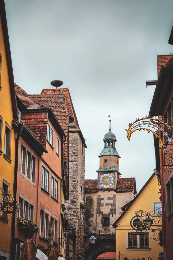A narrow street in a historic European town, flanked by colorful buildings with a clock tower and a stork nest visible in the background.