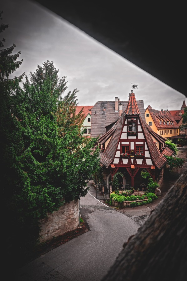 A view of a charming, half-timbered house with a steep roof and dormer windows, set among lush greenery and adjacent to a winding street in the quaint town of Rothenburg, Germany.