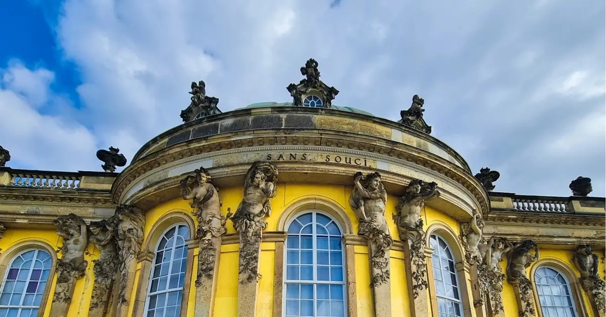 Sanssouci Palace in Potsdam, showing its curved yellow façade with ornate statues and arched windows beneath a dramatic sky.