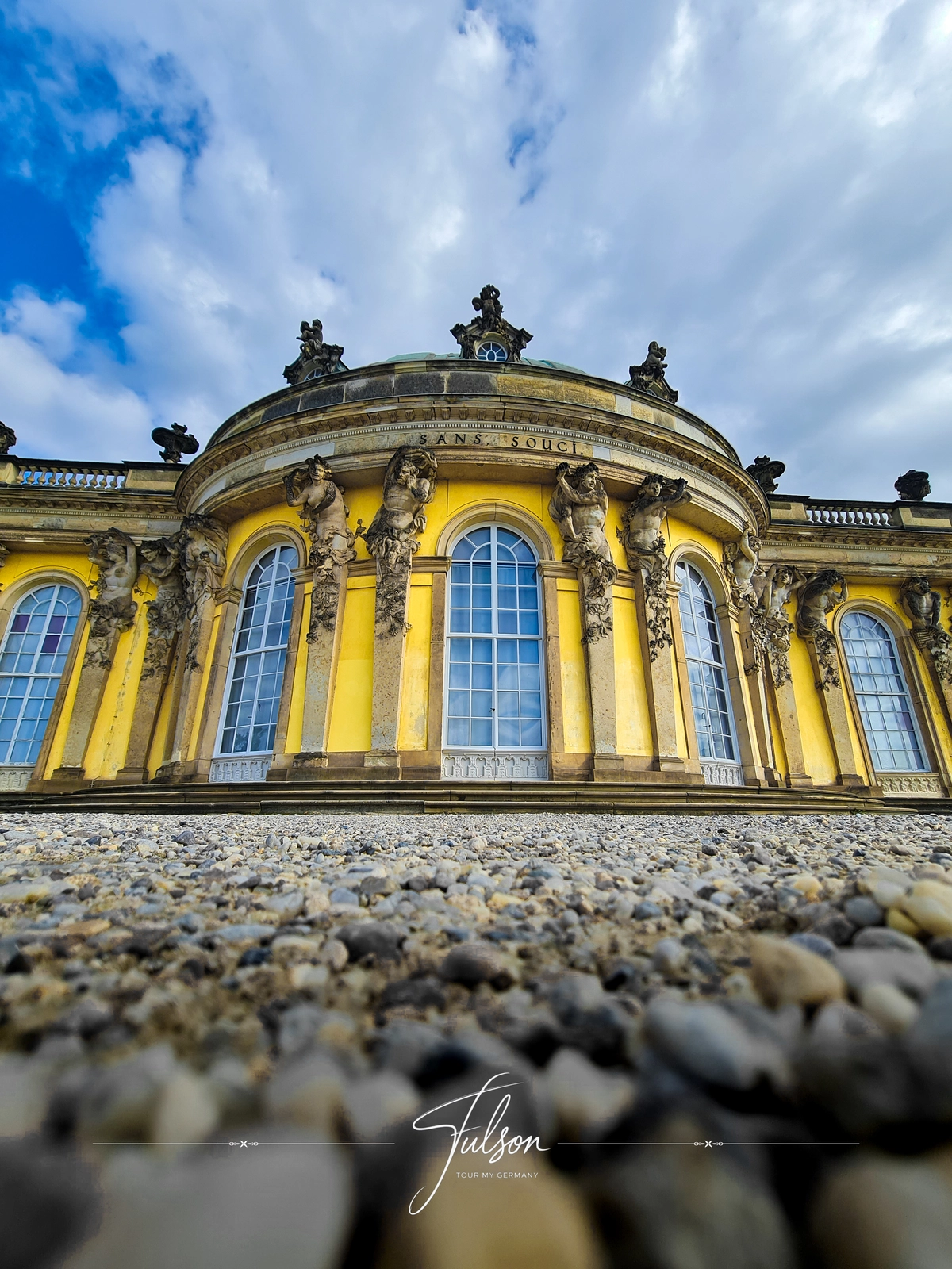 Ornate Sanssouci Palace in Potsdam with yellow building facade with arched windows, statues, and stone details. Photographed from a low angle against a cloudy sky.