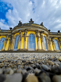 Ornate Sanssouci Palace in Potsdam with yellow building facade with arched windows, statues, and stone details. Photographed from a low angle against a cloudy sky.