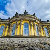 Close-up of Sanssouci Palace in Potsdam, showing its curved yellow façade with ornate statues and arched windows beneath a dramatic sky.