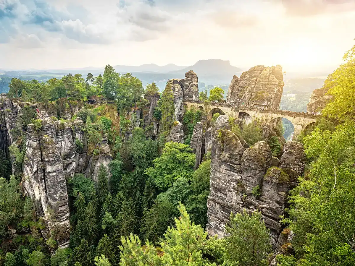 A stone bridge spans between tall, rugged rock formations amidst a forest, with a hazy sky and distant mountains in the background.