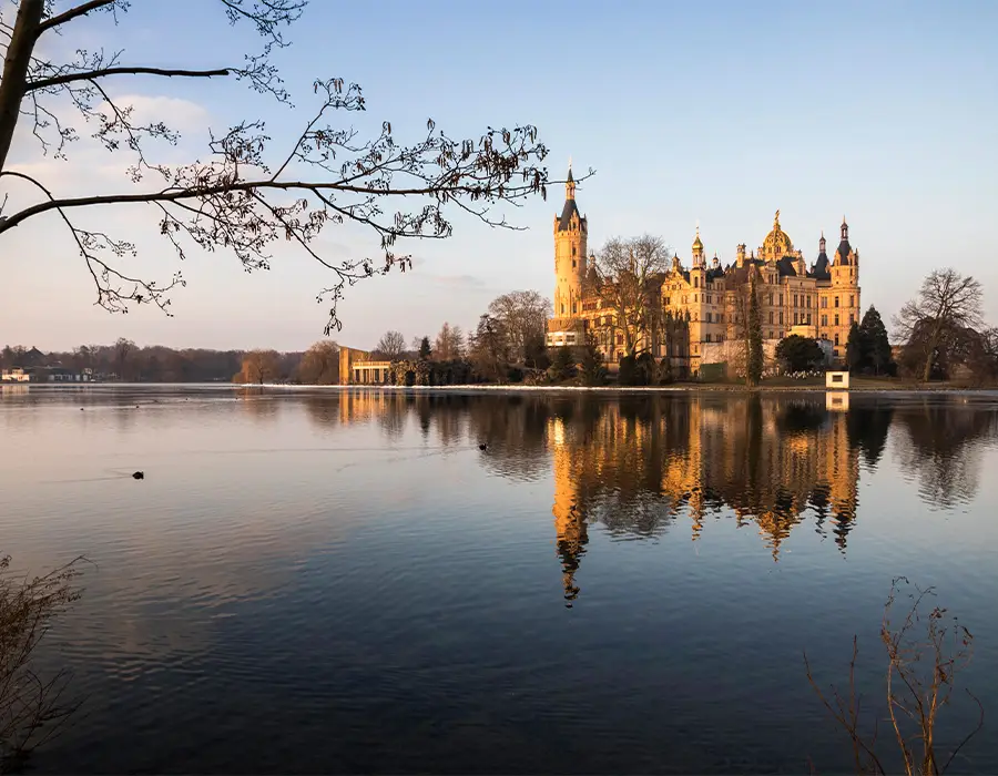 Schwerin Castle in northern Germany reflected in the still lake below, with soft golden light illuminating the towers and autumn trees.