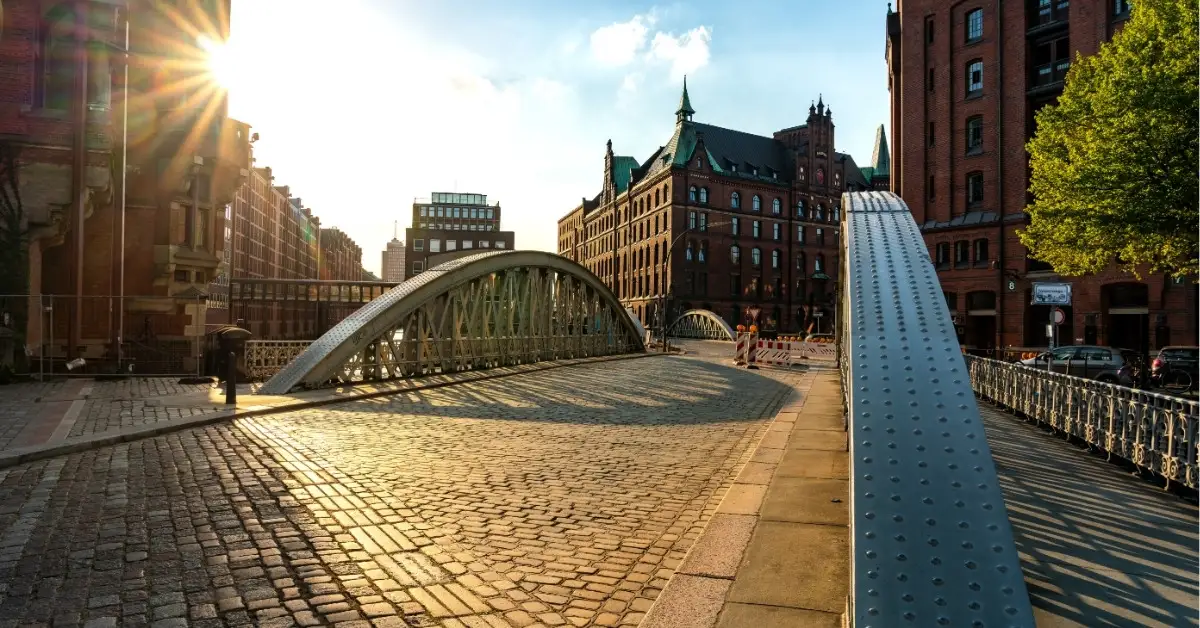 View of a sunlit bridge leading into Hamburg’s Speicherstadt district, with red brick warehouse buildings and cobblestone streets.