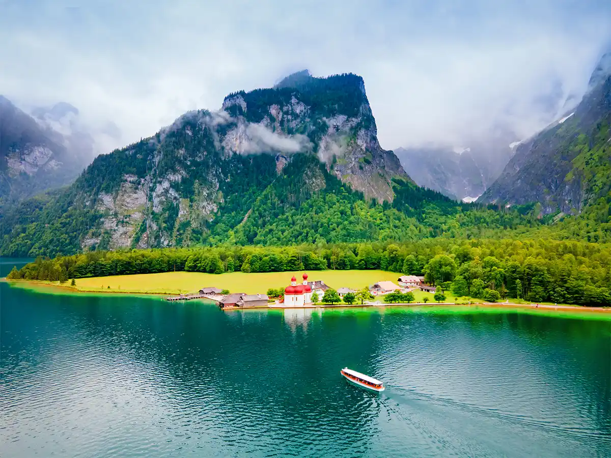 A boat on a calm lake with a red-roofed building on the shore, surrounded by lush greenery and misty Bavarian mountains.