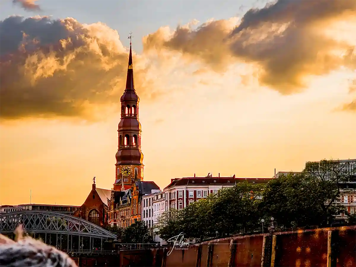 St. Michael's Church in Hamburg with a steeple stands among buildings at sunset, under a partly cloudy sky, with trees and a bridge in the foreground. St. Michael's Church in Hamburg with a steeple stands among buildings at sunset, under a partly cloudy sky, with trees and a bridge in the foreground.