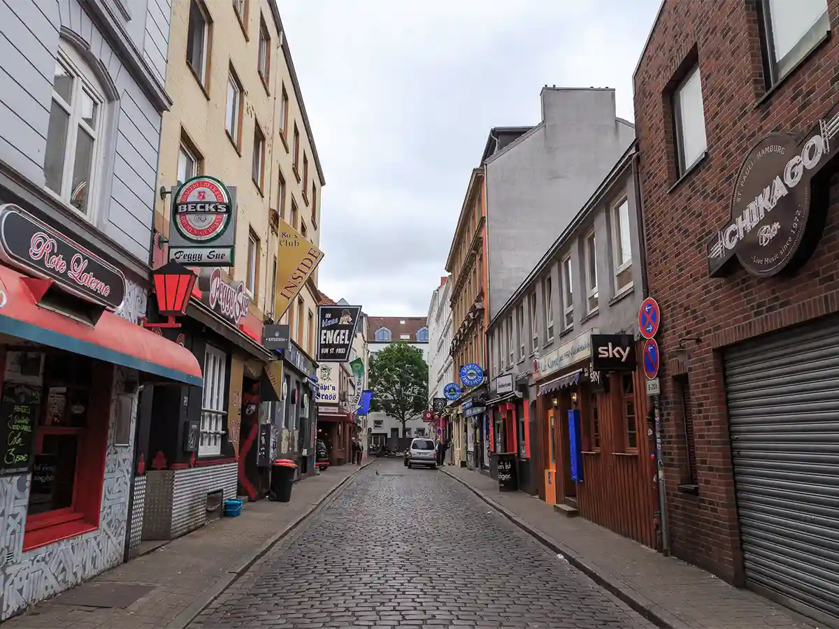 A narrow cobblestone street lined with bars, restaurants, and shops on both sides, with no people present under an overcast sky. A narrow cobblestone street lined with bars, restaurants, and shops on both sides, with no people present under an overcast sky.