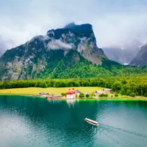 Scenic view of Lake Königssee with a boat approaching St. Bartholomew's Church, set against lush greenery and dramatic alpine cliffs