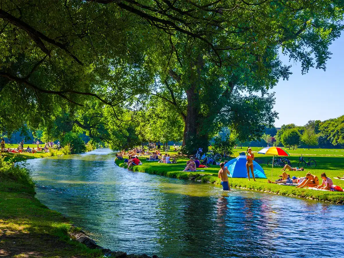 People relaxing by a river on a sunny day, with some sitting under trees and others near colorful umbrellas. A calm stream flows through the park surrounded by lush greenery.