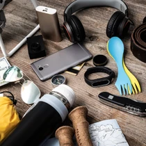 Flat lay of travel essentials including a phone, headphones, spork, map, compass, and power bank on a wooden surface