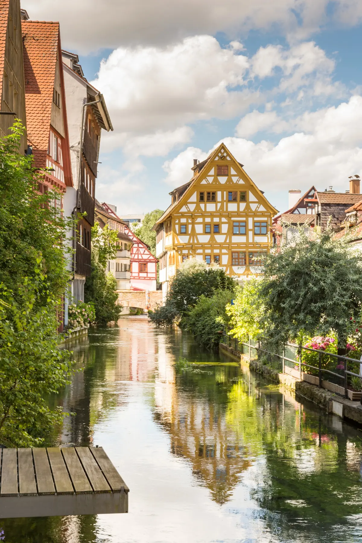 Scenic view of a narrow canal flanked by half-timbered houses and lush greenery under a partly cloudy sky.