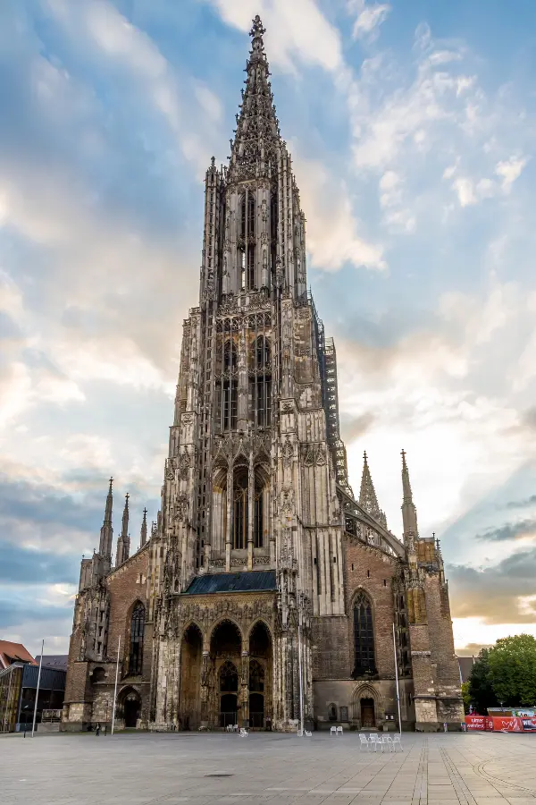 A tall, ornate Gothic cathedral, Ulm Minster, with a detailed facade and spire under a partly cloudy sky.