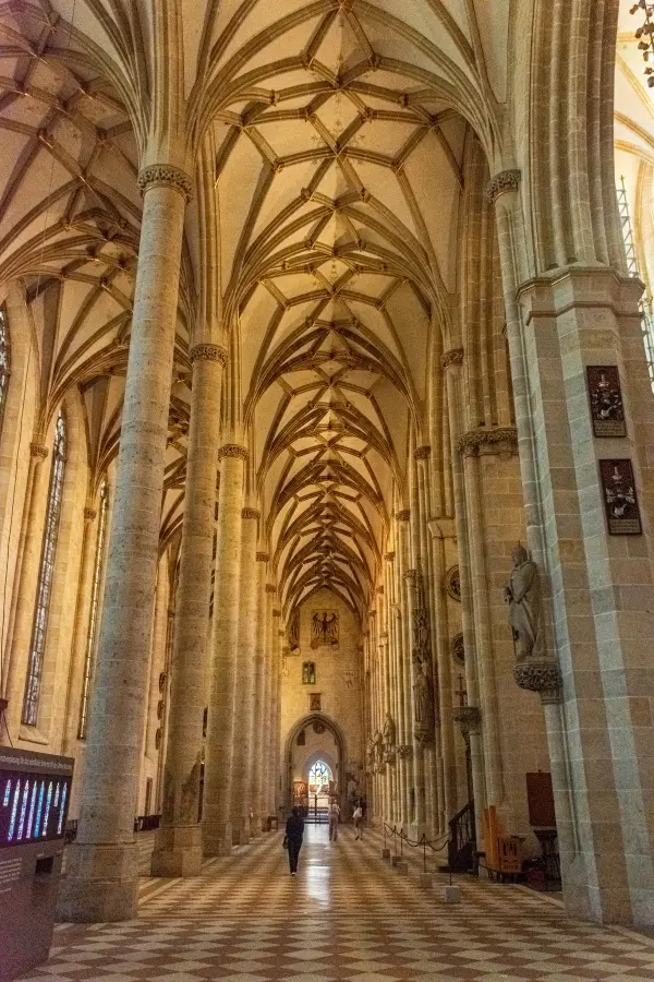 Interior of a grand cathedral with vaulted ceilings, stone columns, and a checkered floor. People walk along the aisle toward a distant entrance.