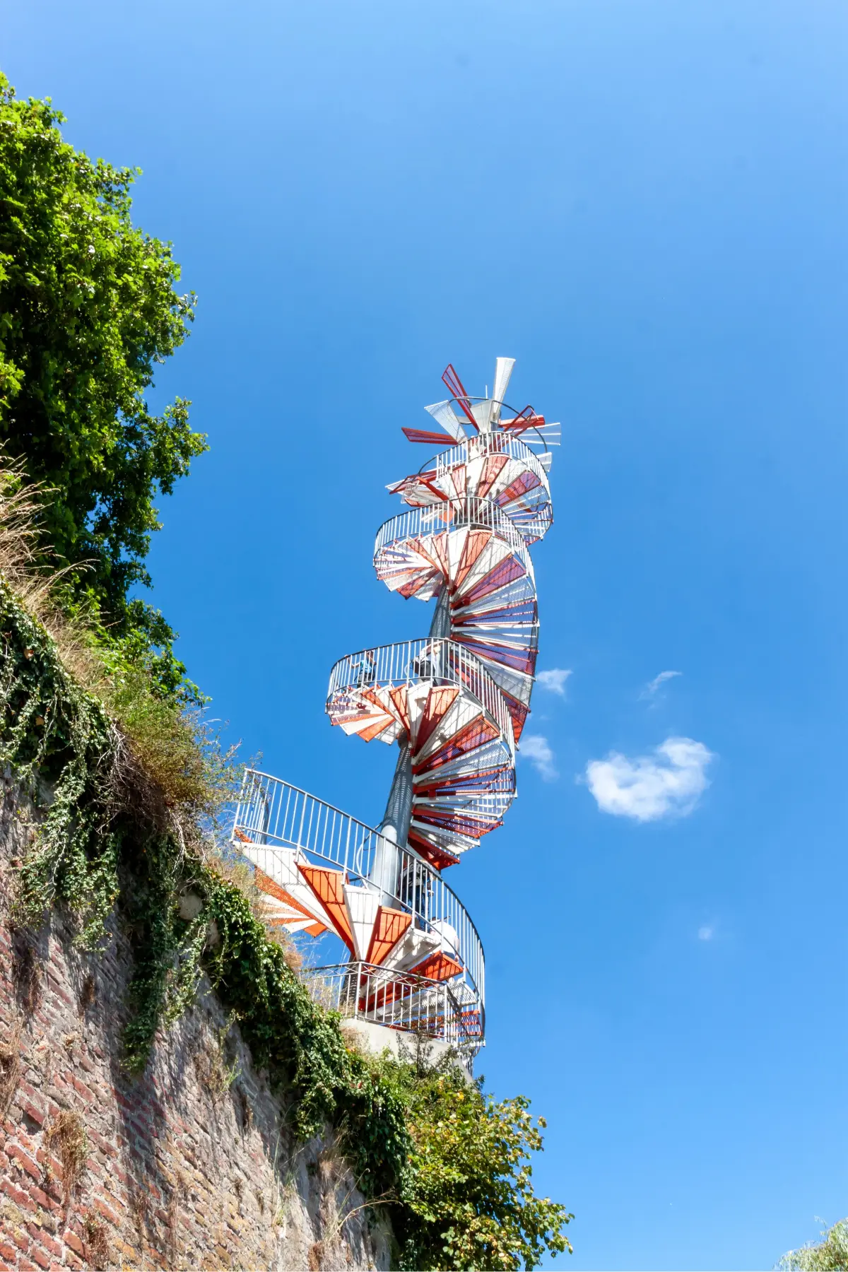 Spiral staircase with red and white steps, extending upward against a bright blue sky, surrounded by greenery and brick wall.