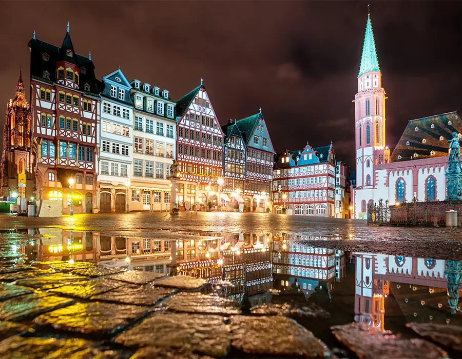 Nighttime view of Römerberg square in Frankfurt, Germany, with half-timbered houses lit up and reflected in rain puddles on cobblestones.