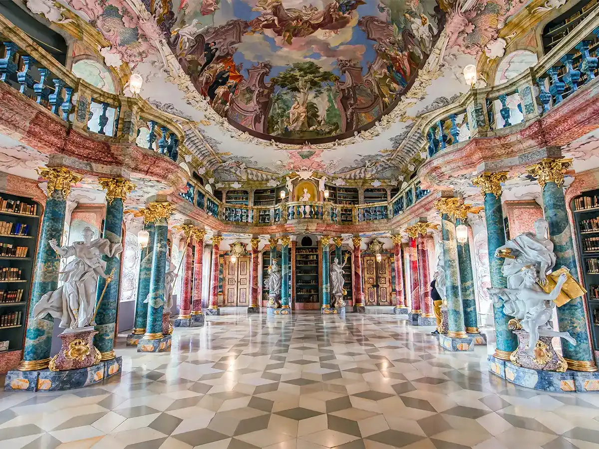 Ornate library interior with colorful murals, marble columns, and statues. Shelves filled with books line the walls under a decorative domed ceiling. Geometric patterned floor.