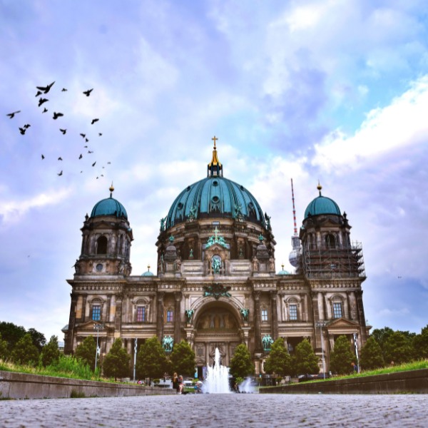 Brown building with green dome-topped roof of the Berlin Cathedral. Birds flying the in sky and fountain in front.