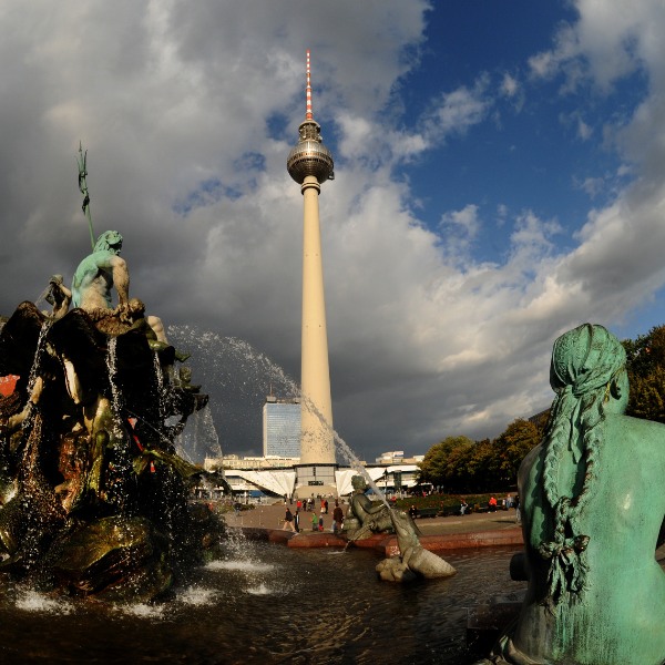 Berlin TV Tower with silver sphere and red and white antennae. Bronze statues around fountain on ground.