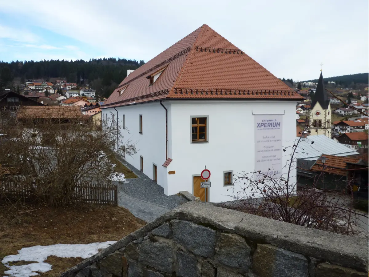 Bayerwald Xperium A white building with a red-tiled roof in a village setting. The banner says "Xperium." A path and stone wall are in the foreground, with a church visible in the background.