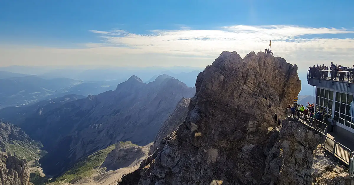 View from the summit of Zugspitze, Germany’s highest mountain, featuring jagged peaks, a golden summit cross, and visitors on an observation deck.