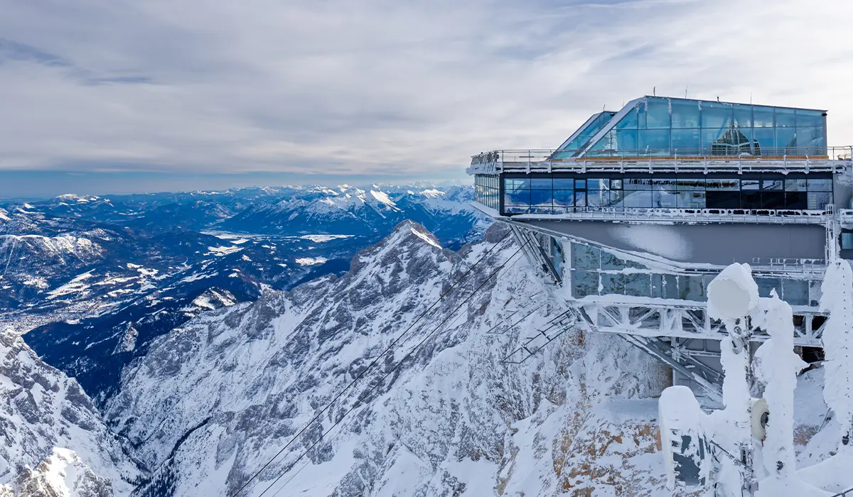 Snow-covered mountain peak with a glass-walled observation deck overlooking a vast, scenic landscape at Zugspitze, Germany.